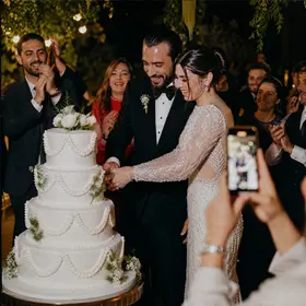 Gelin ve damat düğün pastasını kesiyor; bir misafir telefonla fotoğraf çekiyor